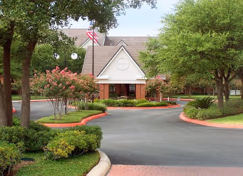 Front entrance and covered porte-cochère of a senior living facility with a circular driveway, landscaped trees, and an American flag.