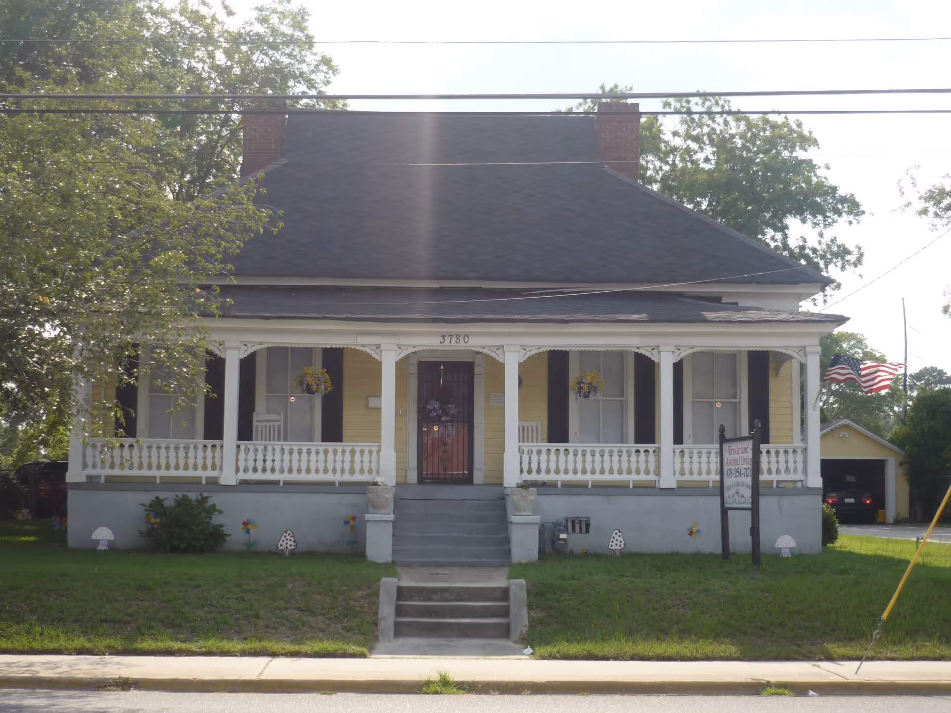 Front exterior view of a yellow house with a large covered porch, white railings, and steps leading up to the front door. There are hanging flower baskets on the porch and an American flag on the right side near a detached garage. The house number 3780 is visible above the door.