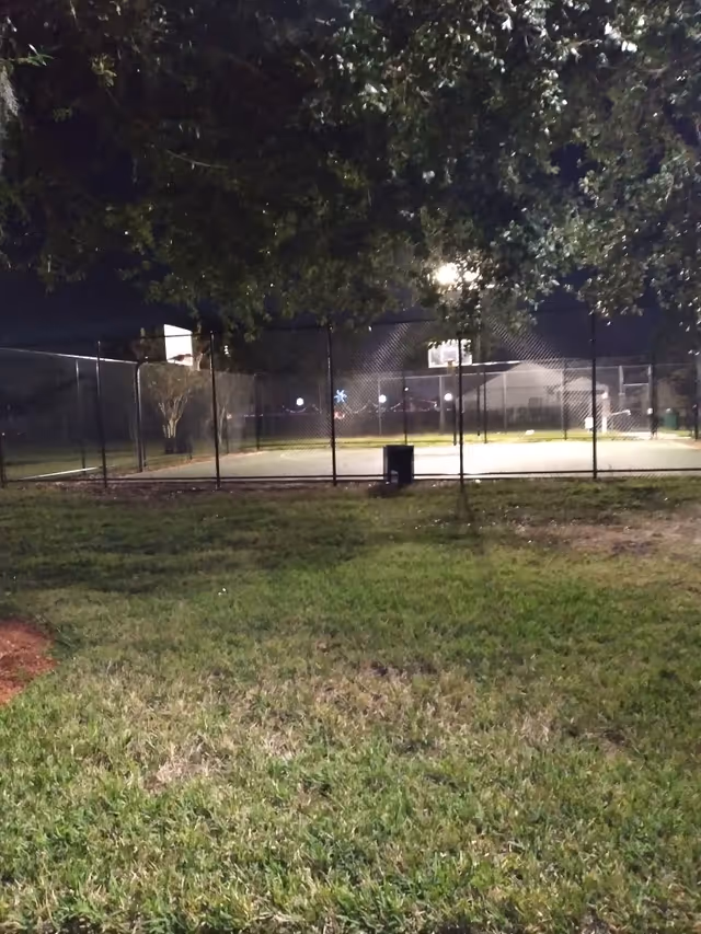 Night view of a fenced outdoor basketball court with grass, trees, and lights.