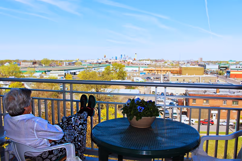 Senior resident relaxing on balcony chair with table and flowers, overlooking city view.