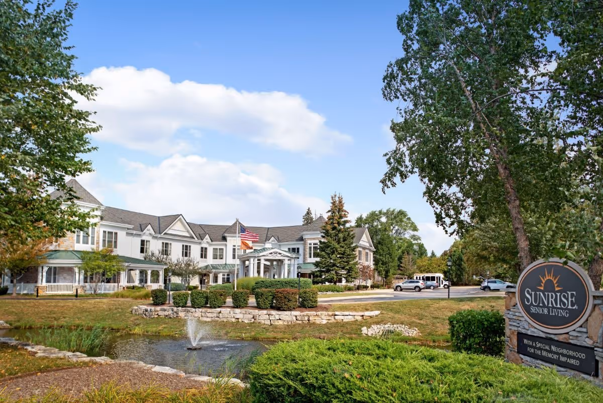Exterior view of a senior living facility building with a pond and fountain in the foreground, surrounded by trees and bushes. A sign on the right reads 'Sunrise Senior Living' with a note about a special neighborhood for the memory impaired. The sky is partly cloudy.