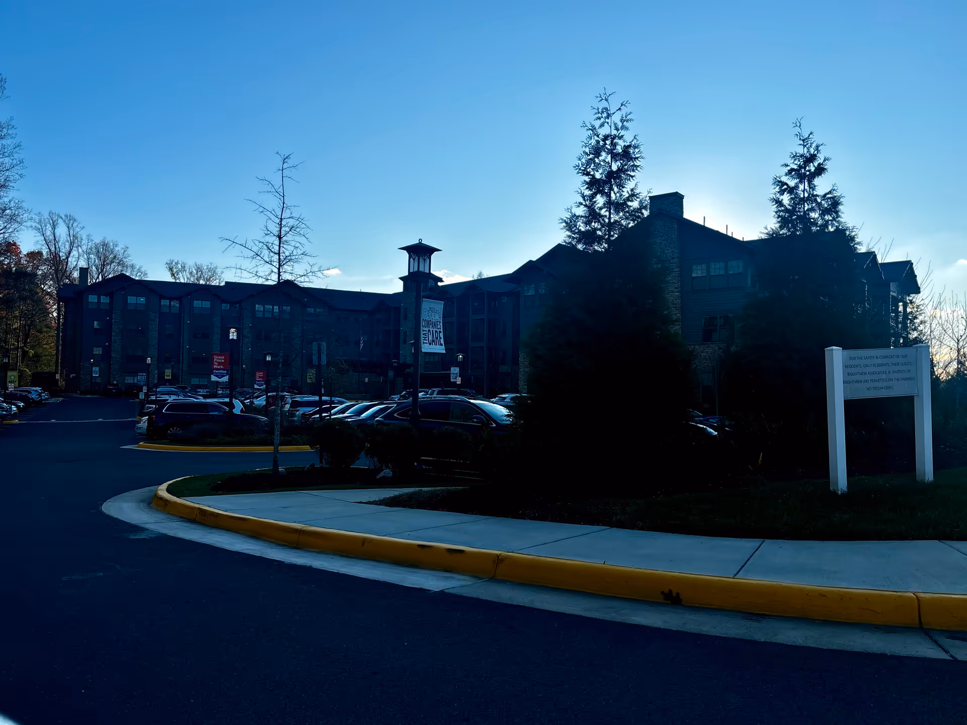 Front exterior of a multi-story senior living building with parked cars, a curved driveway and sidewalk under a clear blue sky.