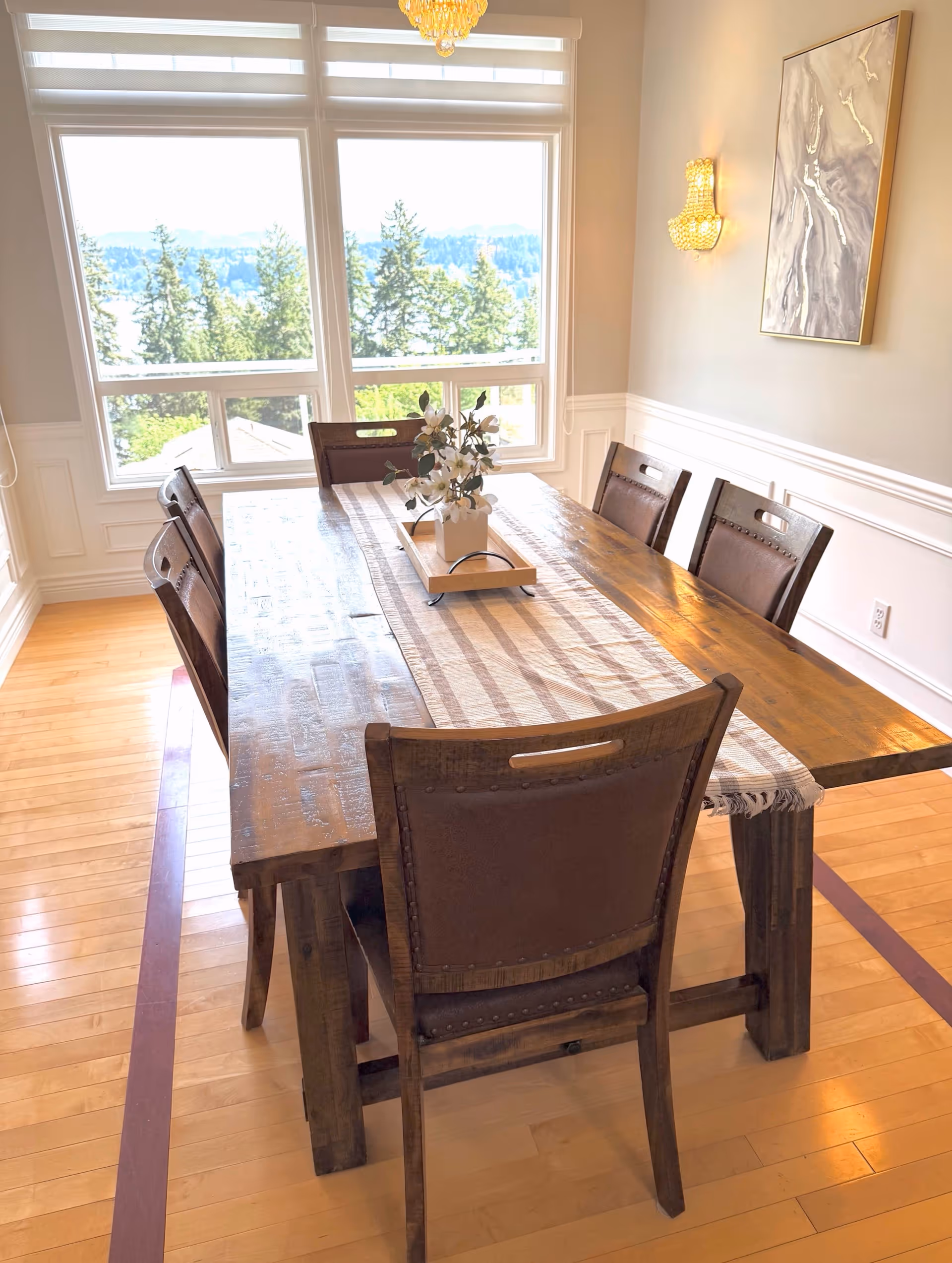 Wooden dining table with chairs in a bright dining room overlooking trees through large windows.