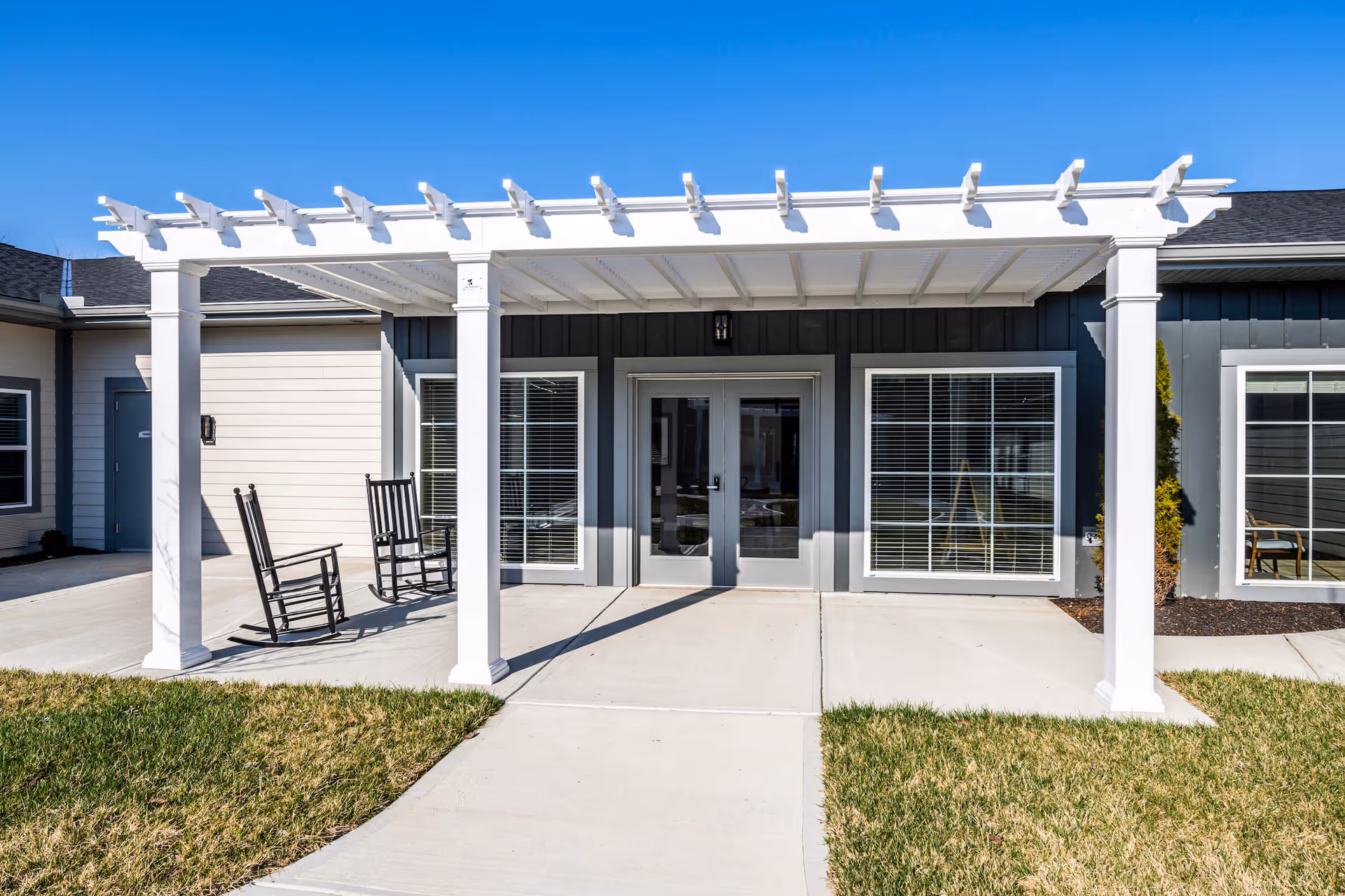 Front entrance of a senior living building with a white pergola, double glass doors, rocking chairs, and large windows.