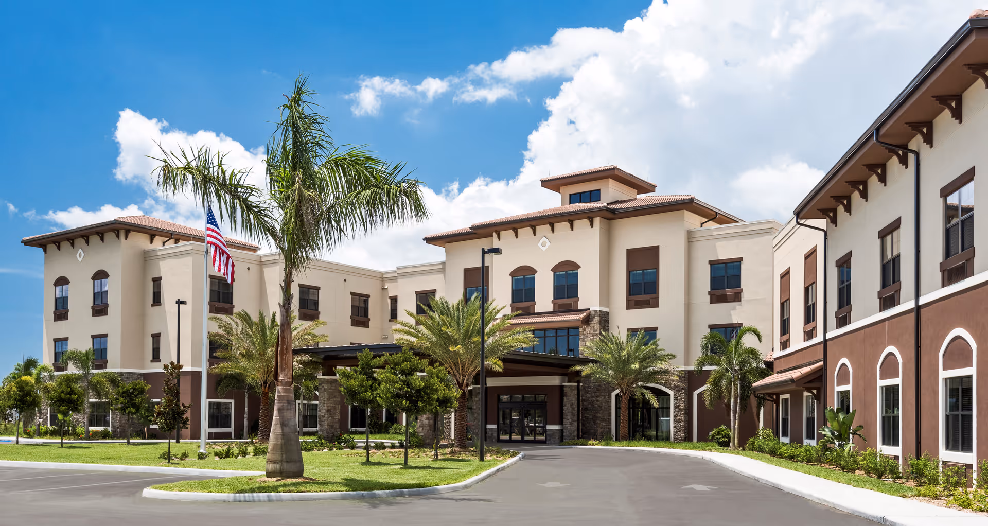 Exterior view of Keystone Place at Beachwalk, a multi-story senior living facility with beige and brown walls, large windows, palm trees, and an American flag in front under a blue sky with scattered clouds.