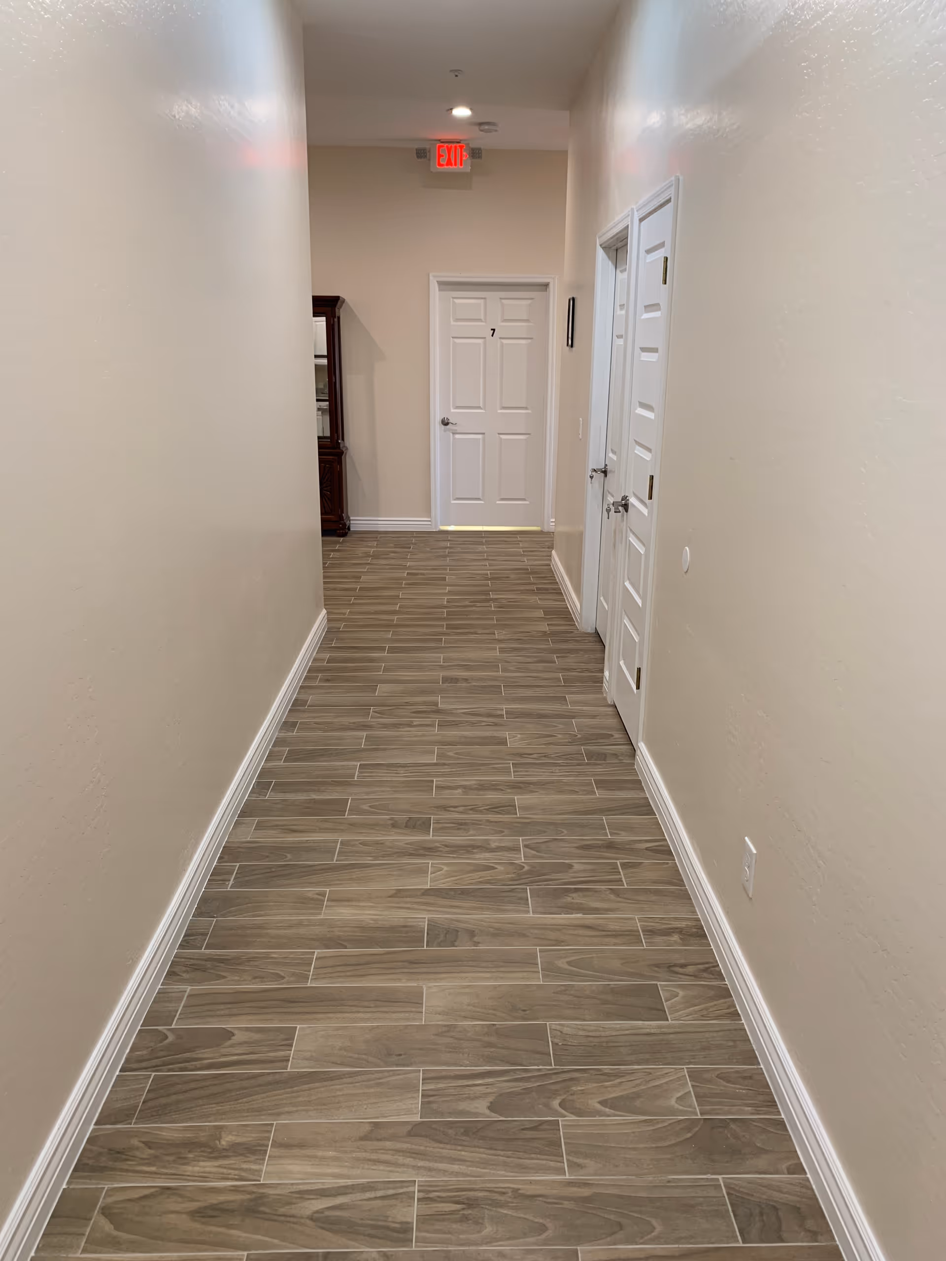 A long, narrow hallway with light beige walls and wood-patterned tile flooring. There are three white doors visible, one at the end of the hallway marked with the number 7, and two on the right side. A red exit sign is mounted above the door at the end. A wooden cabinet is partially visible on the left side near the end of the hallway.