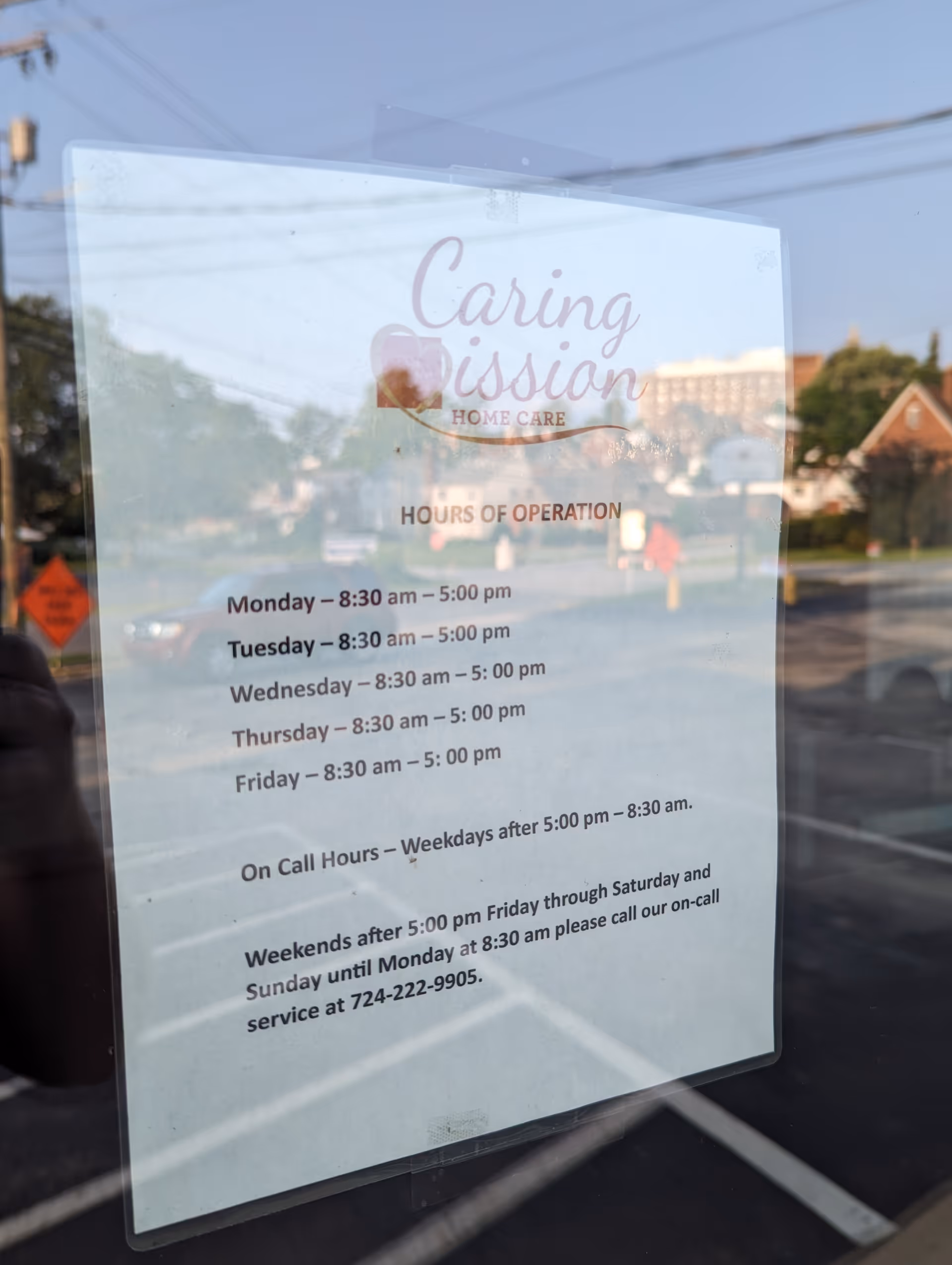 Sign on glass window displaying Caring Mission Home Care hours of operation with street reflection