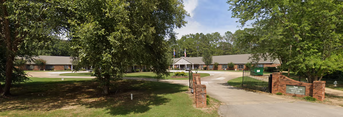 Exterior view of Creekside Manor Assisted Living with driveway, brick sign, and surrounding trees.