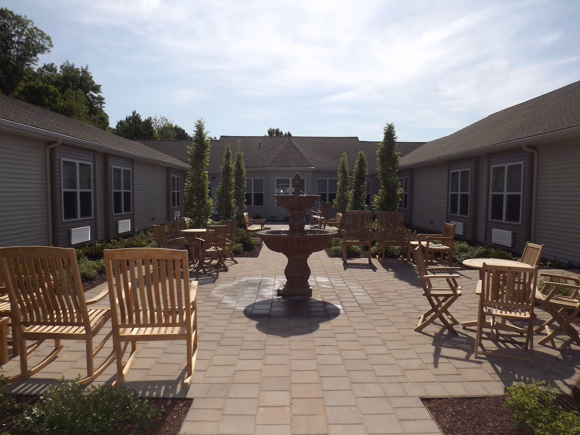 Outdoor courtyard area at Mullica Gardens Assisted Living with a central stone fountain surrounded by wooden chairs and tables. The courtyard is paved with stone tiles and bordered by low bushes and tall, narrow trees. The surrounding building has multiple windows and a sloped roof under a partly cloudy sky.
