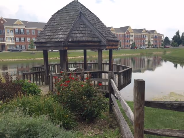 A wooden gazebo with a shingled roof situated by a pond, surrounded by greenery and flowers, with a wooden fence in the foreground and a large multi-story residential building in the background.