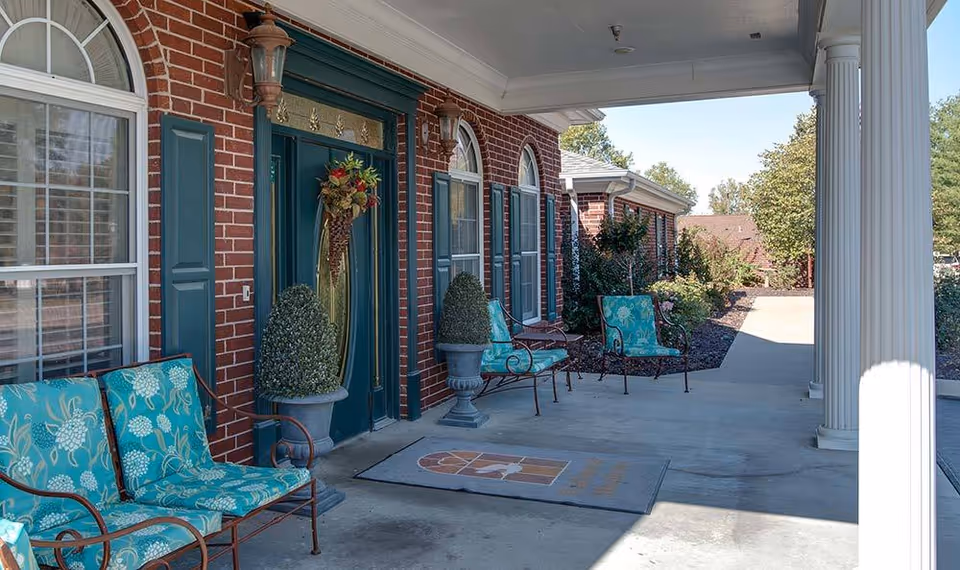 Covered brick entrance with green double doors and wreath, arched windows, columns and turquoise cushioned patio chairs on the porch.
