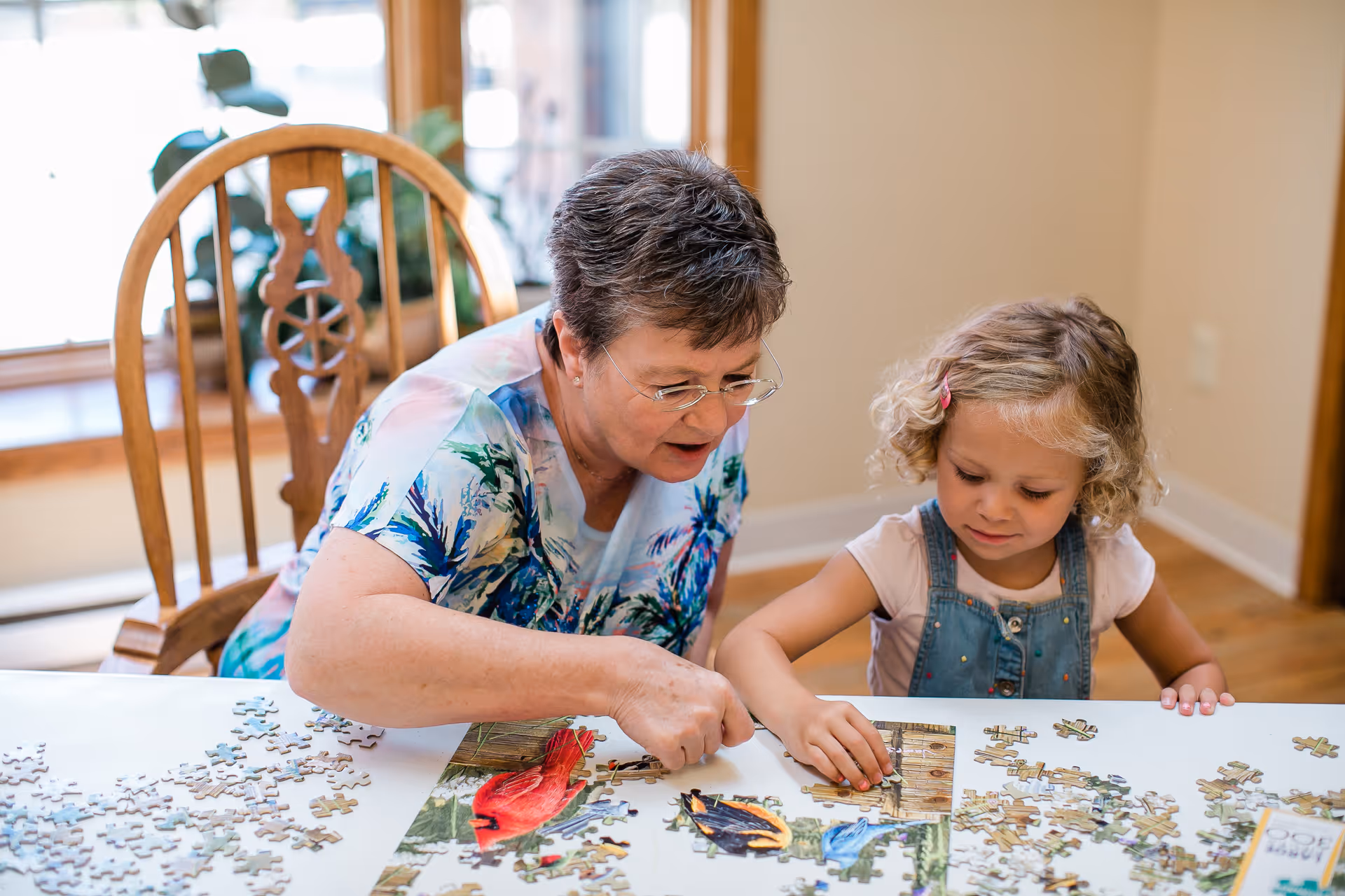 An elderly woman and a young girl sitting at a table working together on assembling a jigsaw puzzle featuring colorful birds. They are in a well-lit room with wooden floors and a wooden chair with a decorative backrest is visible behind them.