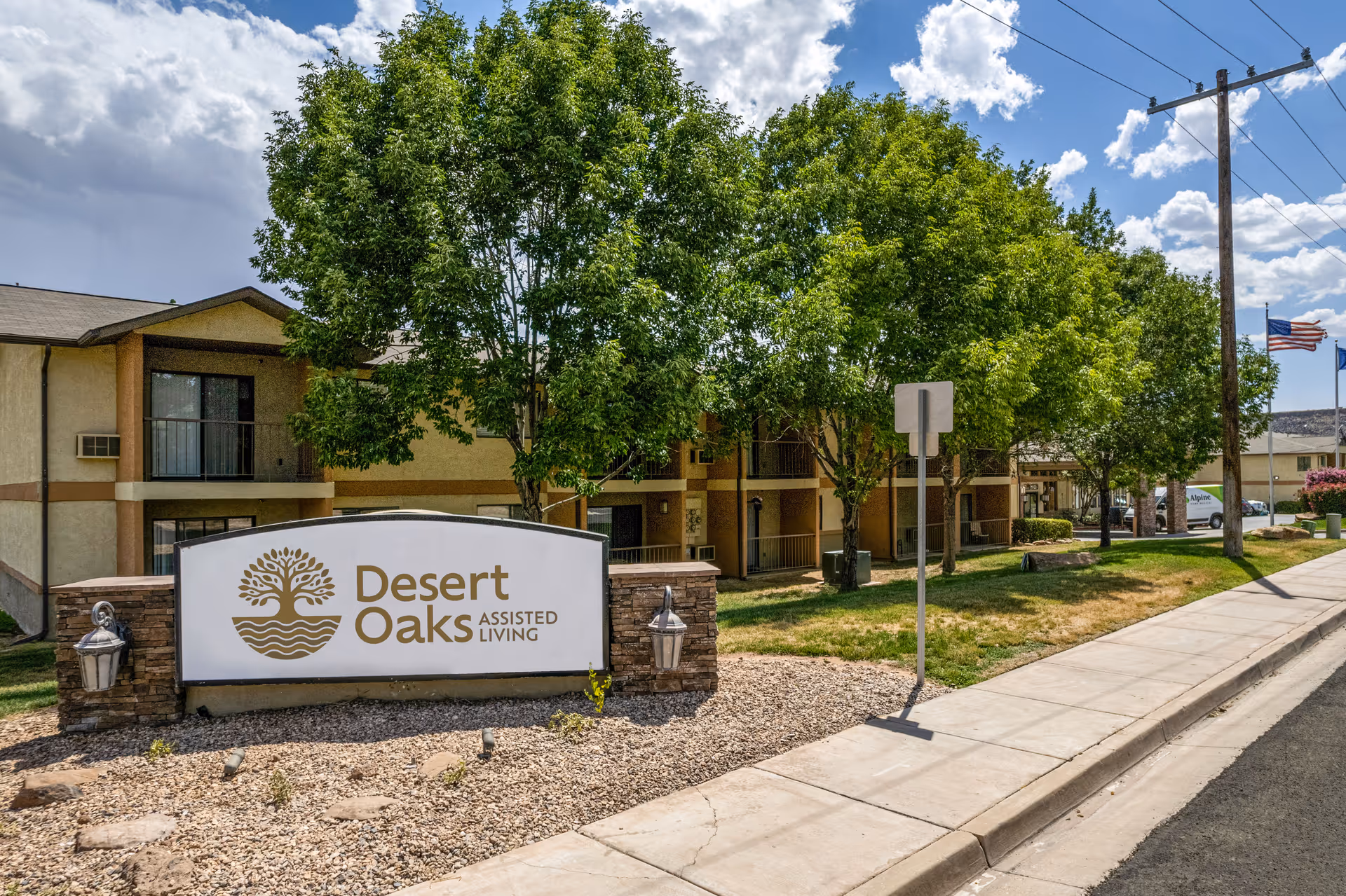 Exterior view of Desert Oaks Assisted Living facility with a large sign in front displaying the facility name and logo. The building is two stories with balconies, surrounded by green trees and a sidewalk along the street. The sky is partly cloudy.