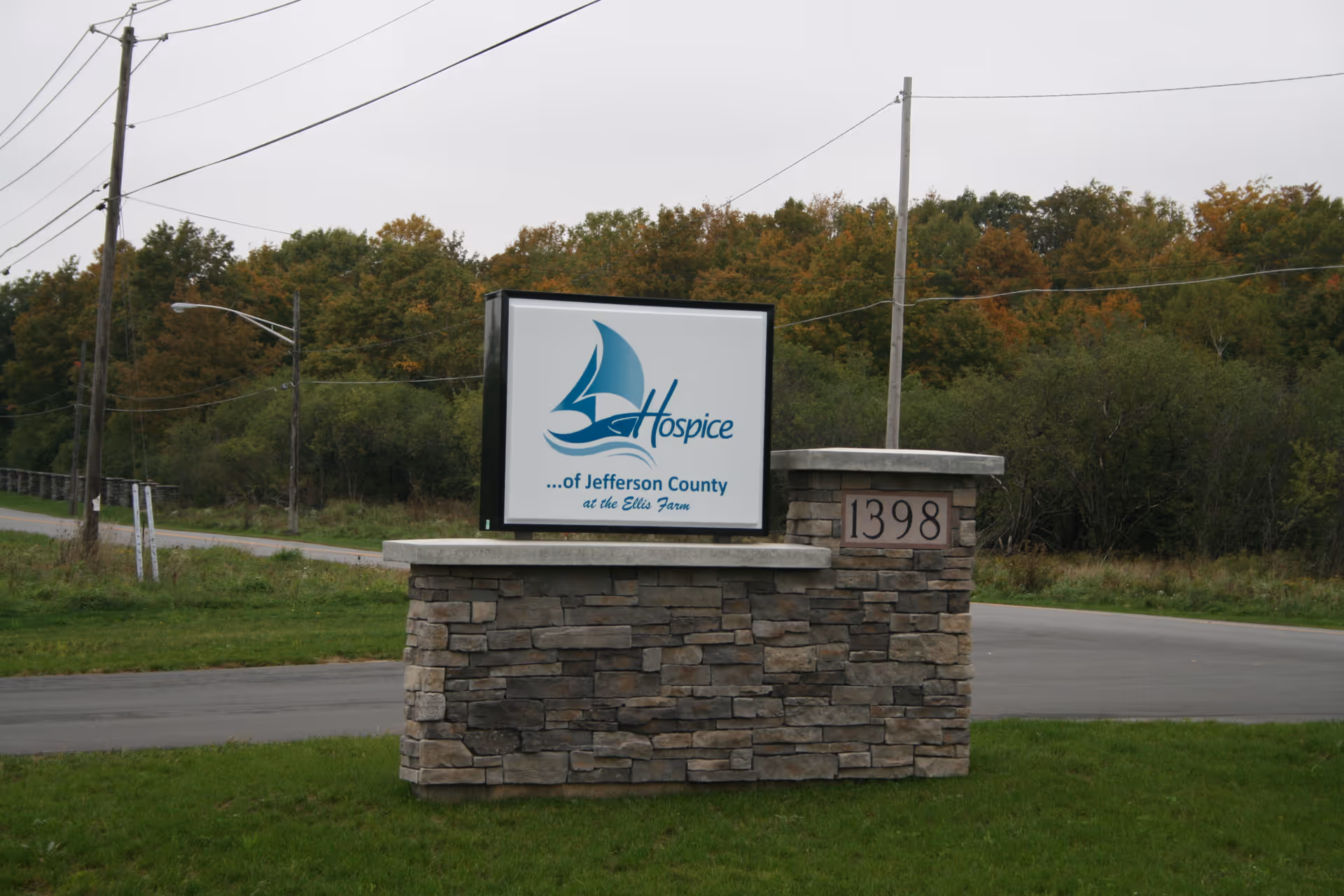 Stone entrance sign for Hospice of Jefferson County beside a road, with fall trees in background.