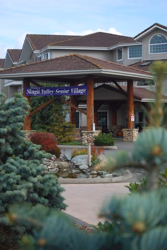 Entrance of a senior living community with a covered driveway supported by wooden pillars and stone bases. A purple banner reads 'Welcome to Skagit Valley Senior Village'. The building has a tiled roof and multiple windows, surrounded by landscaped greenery including bushes and trees.