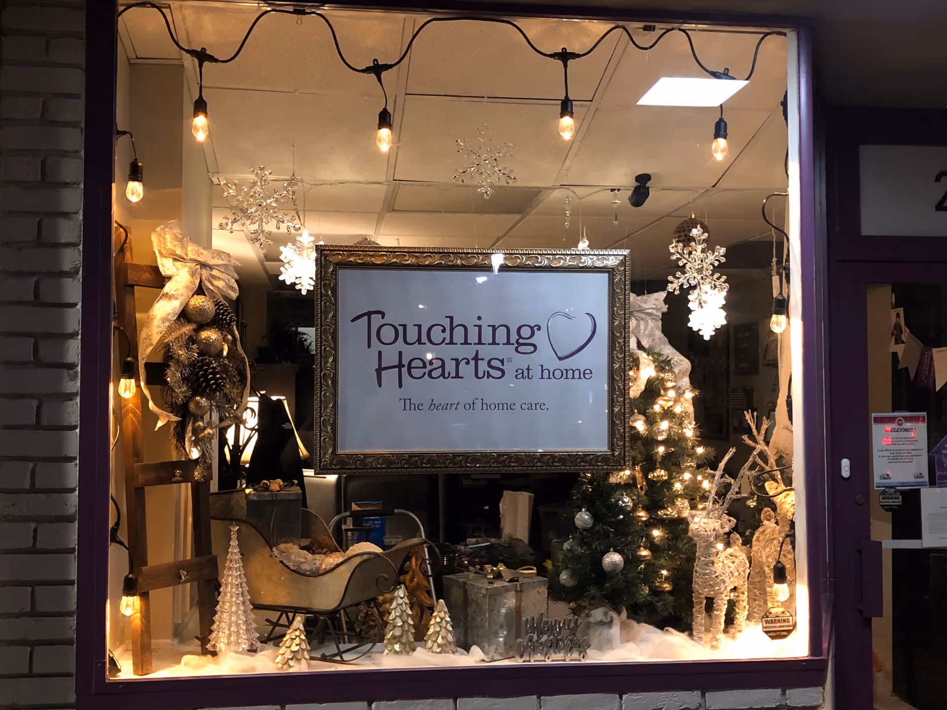 Storefront window display featuring a framed Touching Hearts at home sign and holiday decor.