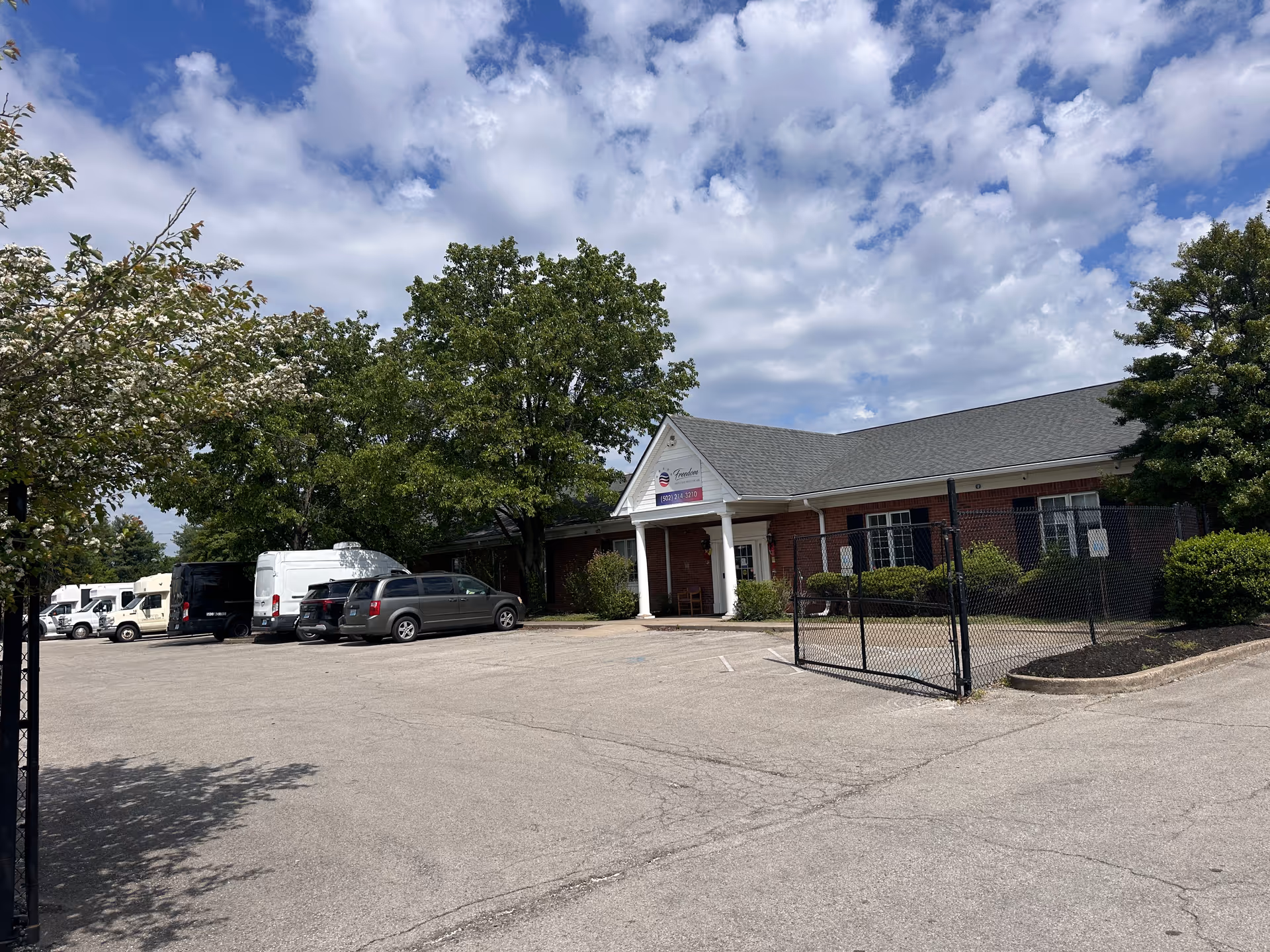 Brick senior living facility with a parking lot, gated entrance, and trees under a blue sky.