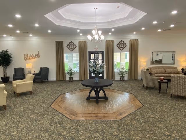 A spacious, well-lit living room area with a central round table holding a vase of flowers on a wooden octagonal platform. The room features patterned carpet, beige and gray armchairs, a sofa, side tables with lamps, decorative wall art, and large windows with curtains. A chandelier hangs from a tray ceiling.