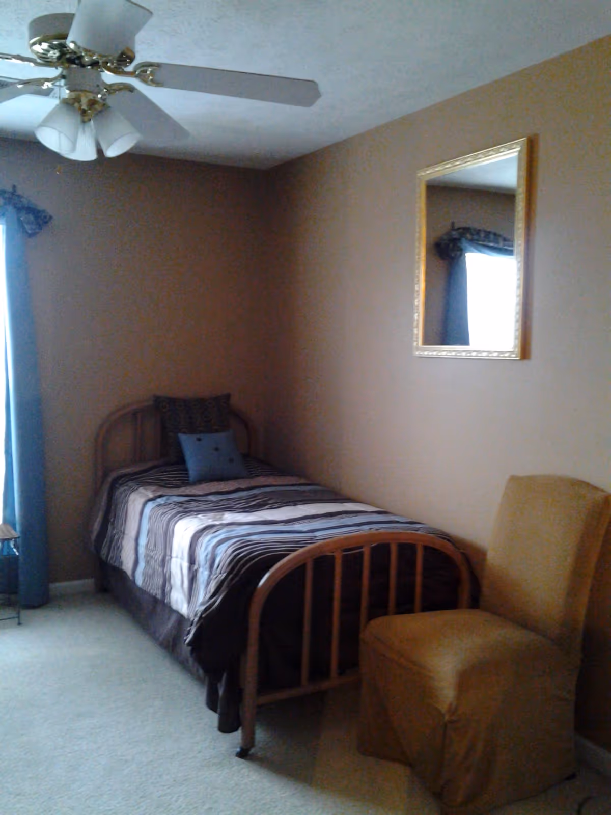 A small bedroom with a single bed, striped bedding, blue curtains, a chair, and wall mirror.