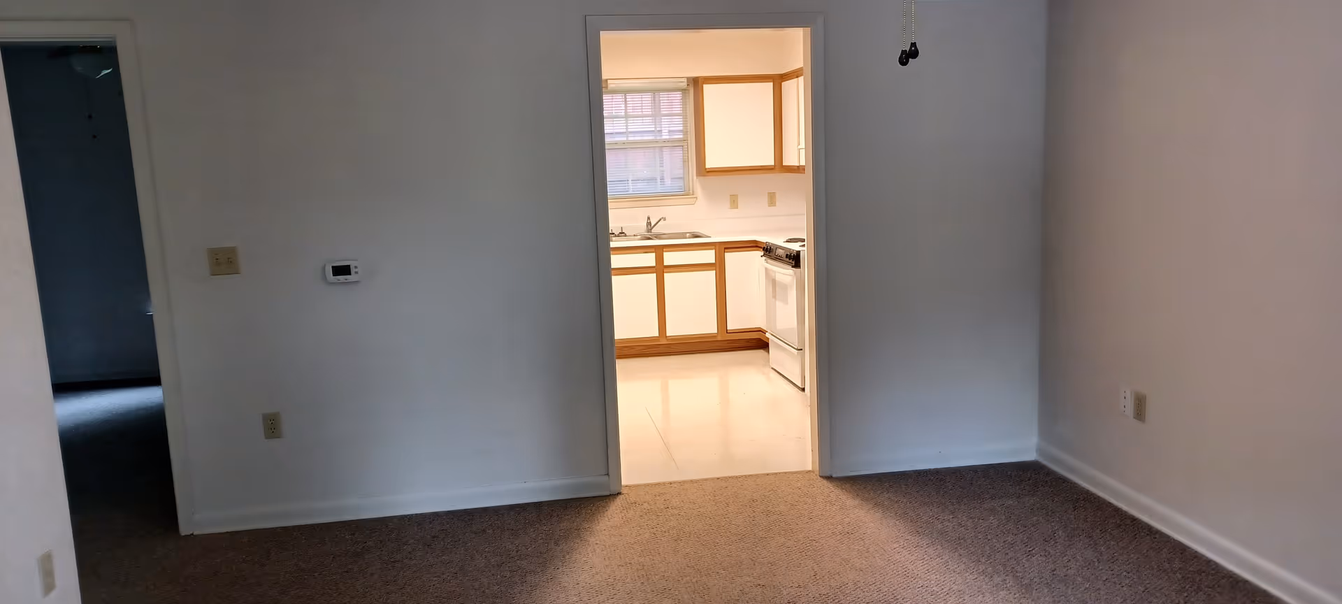 Empty living room area with doorway leading to a bright kitchen.