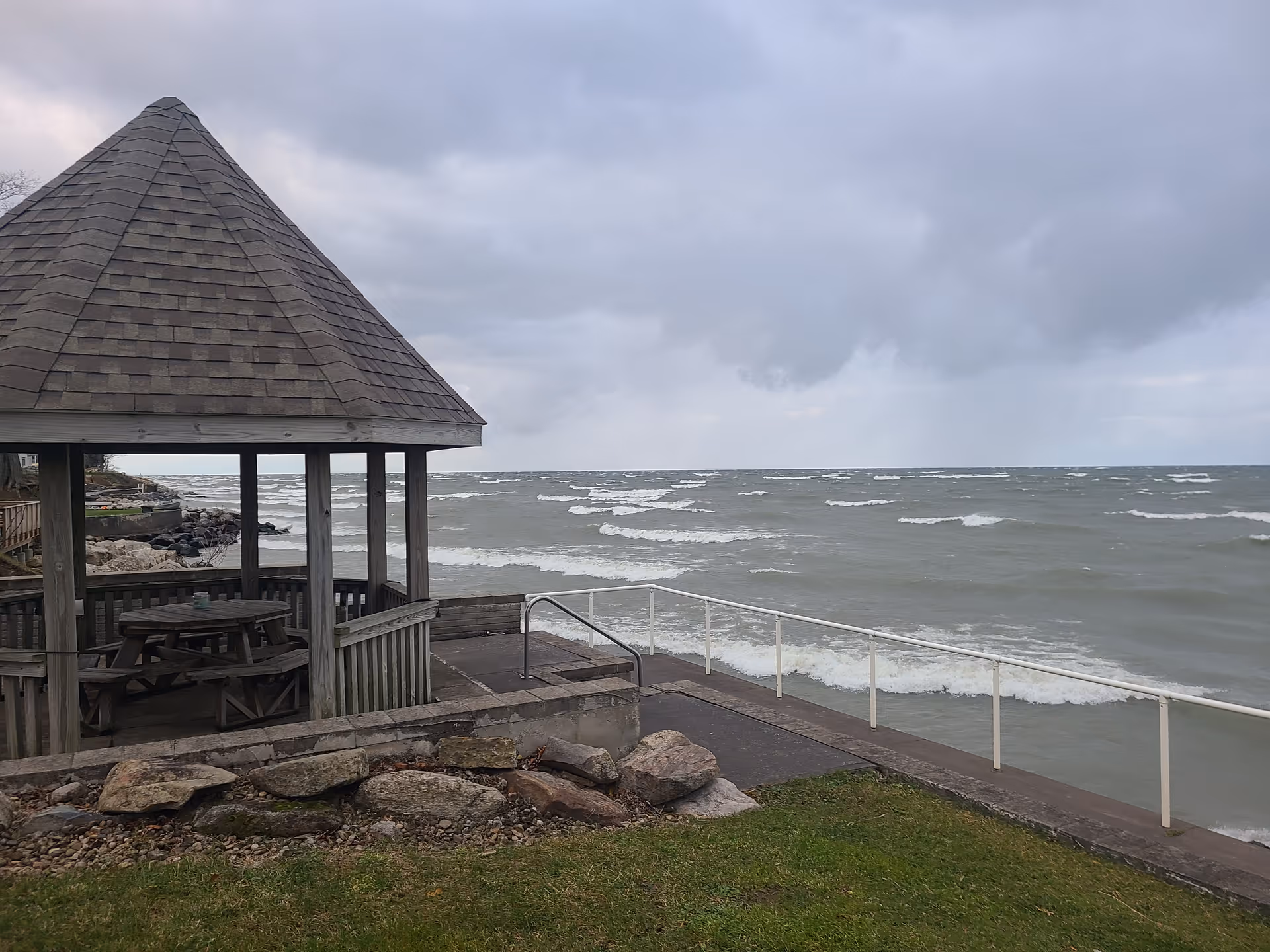 Wooden gazebo with picnic table overlooks a choppy lake and rocky shoreline.