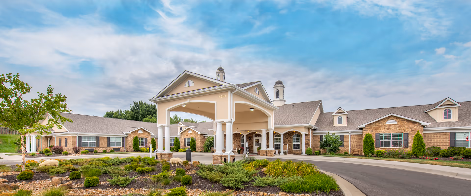 Front exterior view of Benton House of Raymore, a single-story brick building with a covered entrance, surrounded by landscaped greenery and a curved driveway under a partly cloudy sky.