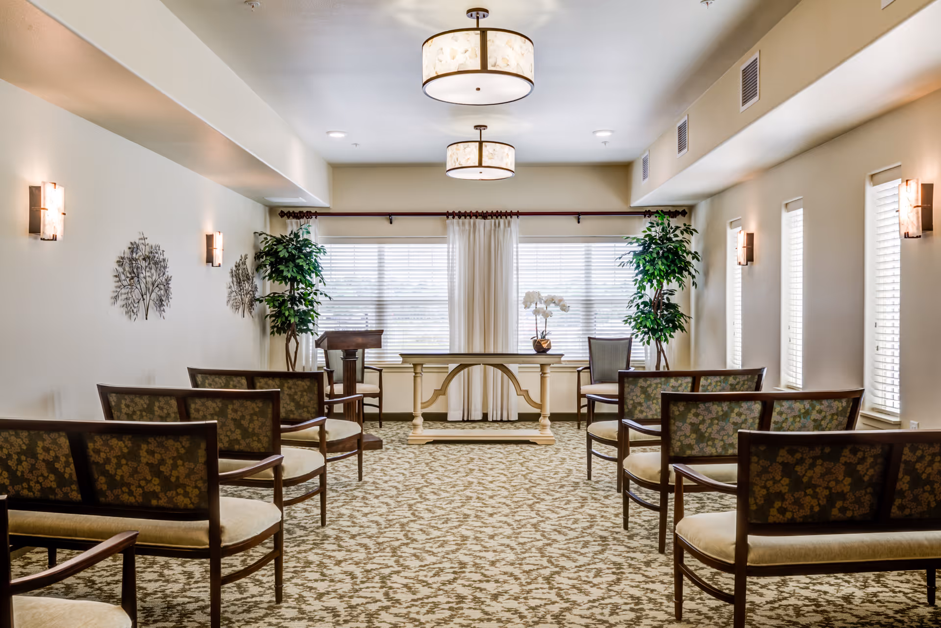 A well-lit room with rows of wooden chairs featuring floral upholstery arranged facing a decorative table and a lectern near large windows with white blinds and curtains. The room has beige walls adorned with metal tree wall art, two large potted plants, patterned carpet, and ceiling light fixtures.