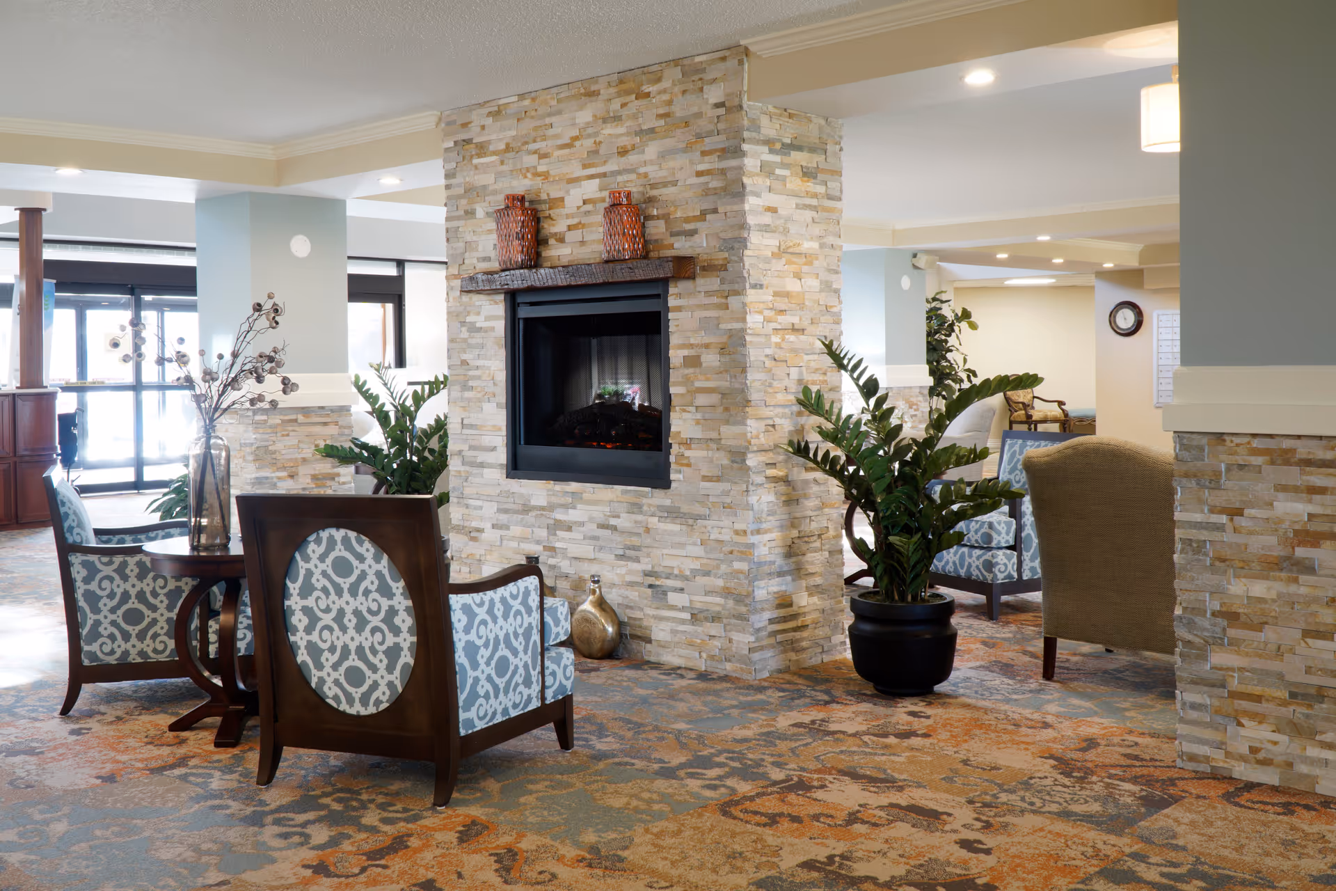 A cozy seating area in a retirement community featuring a stone fireplace in the center, surrounded by patterned armchairs and potted plants. The space has a warm, inviting atmosphere with soft lighting and a decorative carpet.
