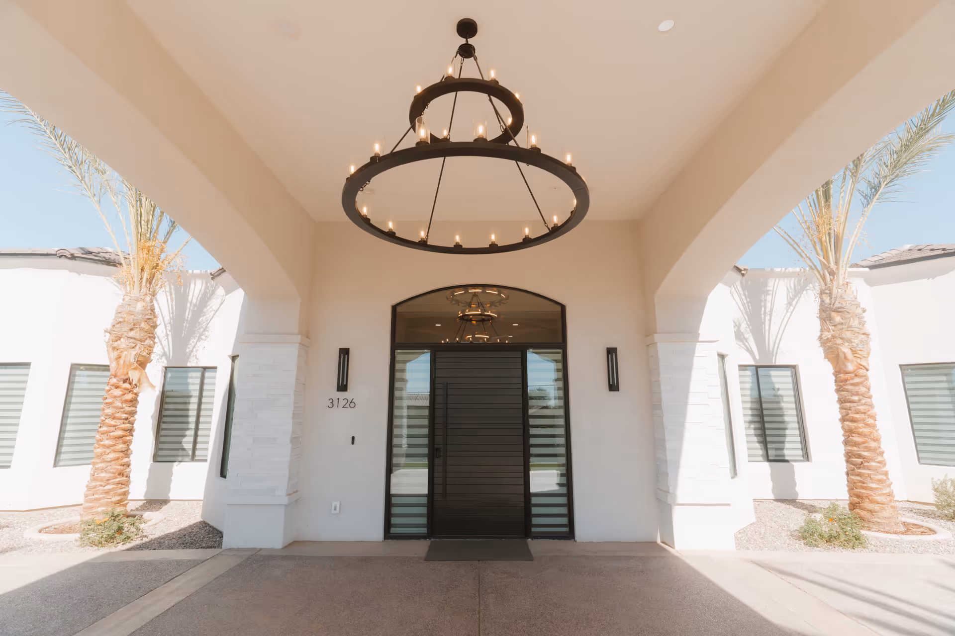 White building entrance with a circular chandelier, glass doors, and palm trees framing the front.