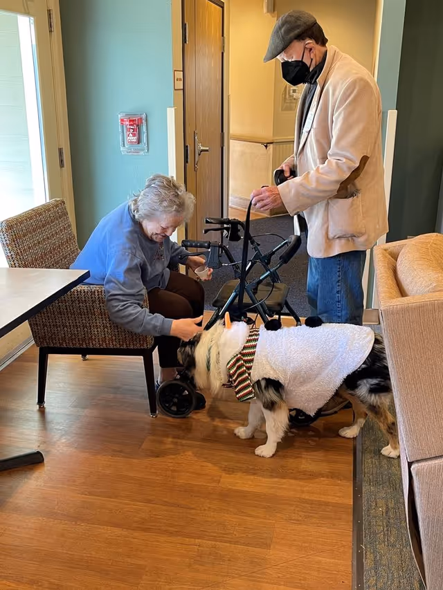 An elderly woman sitting in a chair inside a facility, smiling and petting a dog dressed in a white outfit with a colorful collar. A man wearing a beige jacket, blue jeans, a gray cap, and a black face mask stands nearby holding a walker and the dog's leash. The setting appears to be a common area with wooden flooring and light-colored walls.
