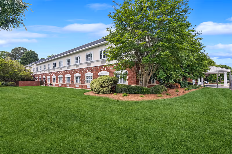 Two-story brick senior living building with a manicured lawn, trees, and a covered entryway.