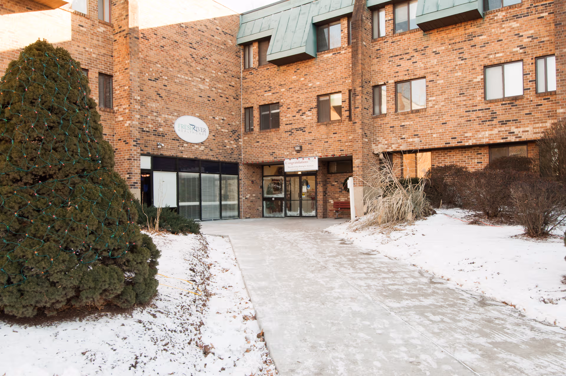 Front entrance of Fresh River Healthcare, LLC building with snowy path and evergreen tree.