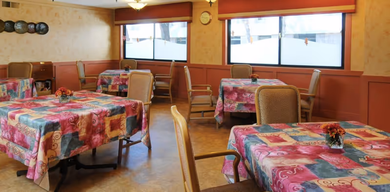 A dining room with several tables covered in colorful patterned tablecloths. Each table has a small flower arrangement in the center. The room has large windows with red valances, beige walls with a subtle pattern, and cushioned chairs around the tables.