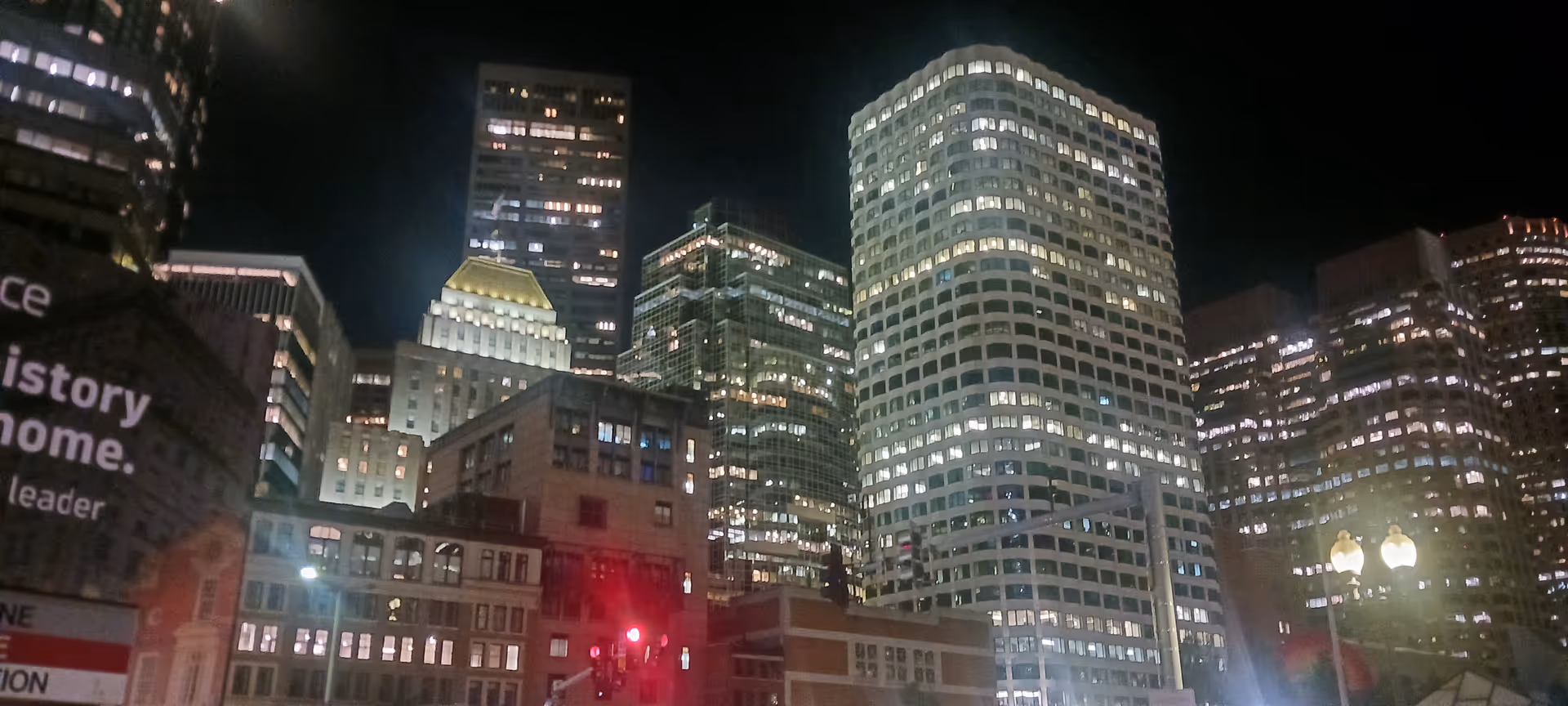 Nighttime city skyline with illuminated skyscrapers and a red traffic light at street level.