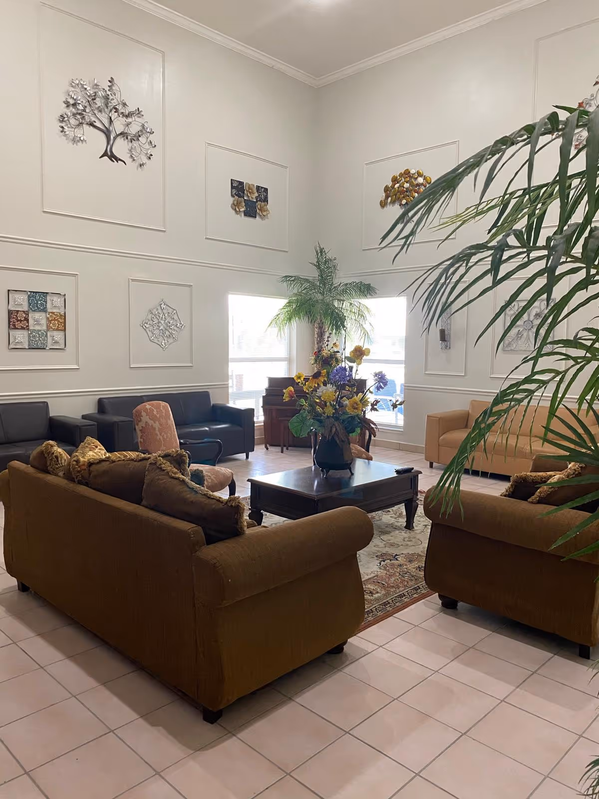 A spacious senior living lounge with brown sofas, a central coffee table, plants, and wall art.