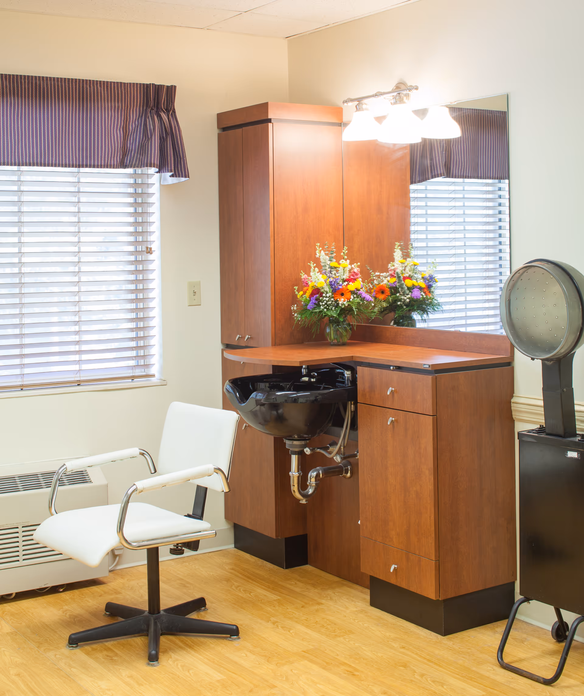 A hair-salon corner with a white styling chair, black shampoo bowl set in wooden cabinetry, mirror, floral arrangement, and a hooded dryer.