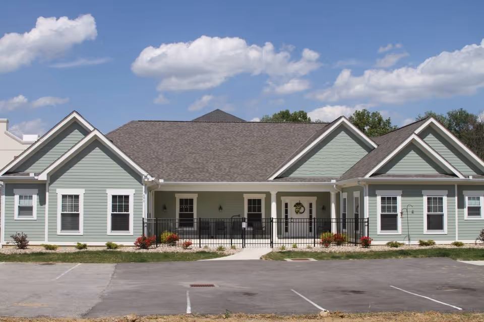 Single-story green senior living facility with a gabled roof, front entrance, and landscaping.