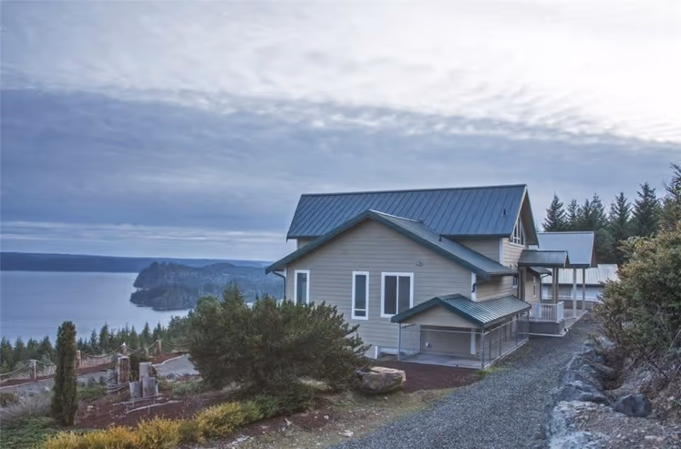 Exterior view of a two-story house with a metal roof situated on a hillside overlooking a large body of water and forested landscape under a cloudy sky.