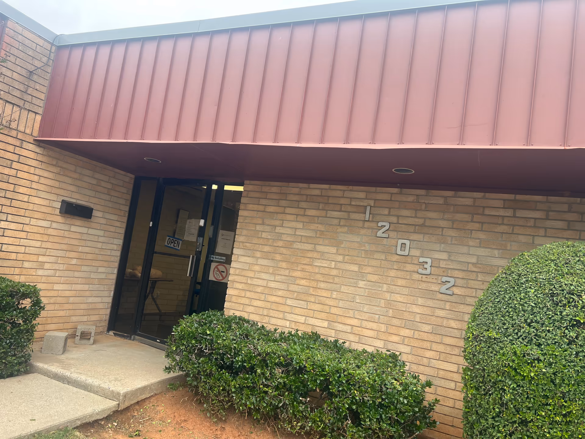 Brick building entrance with a glass door, numbers 2032 on the wall, and bushes nearby.