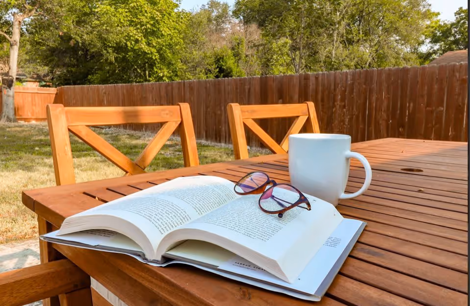 A wooden outdoor table and chairs in a fenced backyard with an open book, reading glasses, and a white mug on the table.