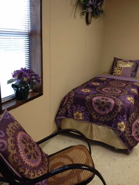 A small bedroom with a single bed covered in a purple patterned bedspread and matching pillow. Next to the bed is a chair with a cushion that has a similar purple pattern. A window with blinds and a small potted plant with purple flowers is on the left wall. A floral wall decoration hangs above the bed.