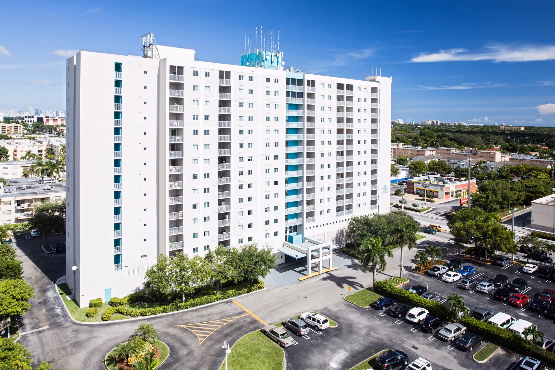 A tall white multi-story residential building with balconies, a driveway and parked cars surrounded by palm trees under a blue sky.