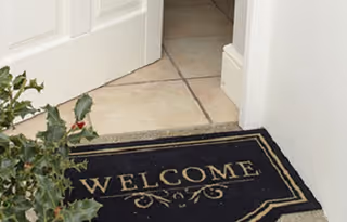 Close-up view of a black welcome mat with gold lettering placed at the entrance of a room with tiled flooring and a white door partially open, next to a green leafy plant.