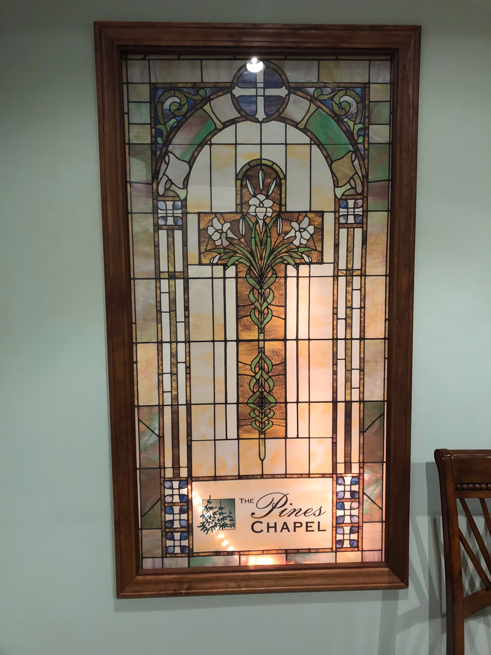 Stained glass window featuring a cross with floral designs and the text 'The Pines Chapel' at the bottom, framed in wood and mounted on a light green wall next to a wooden chair.