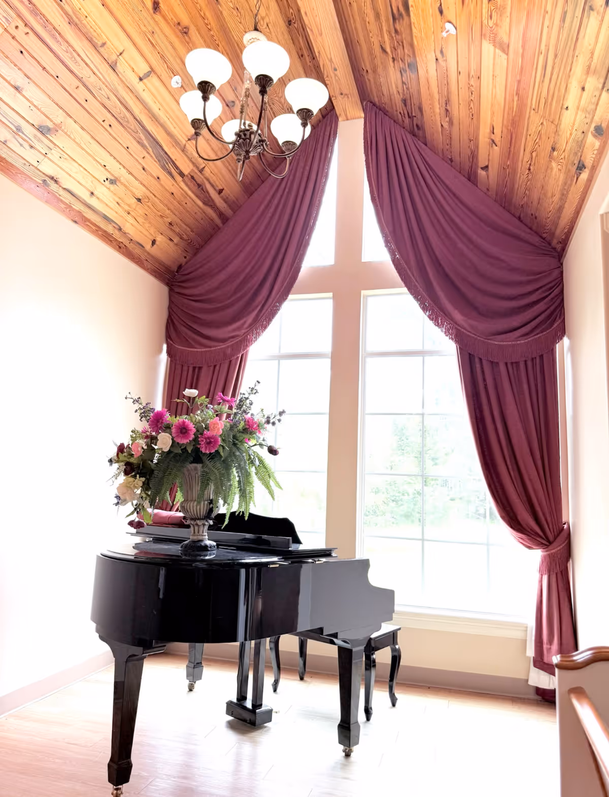 A black grand piano topped with a floral arrangement sits in front of tall windows framed by burgundy drapes beneath a wood-paneled vaulted ceiling and chandelier.