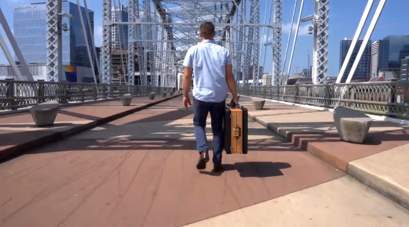 A man walks on a steel bridge walkway, carrying a brown suitcase, with city buildings behind.