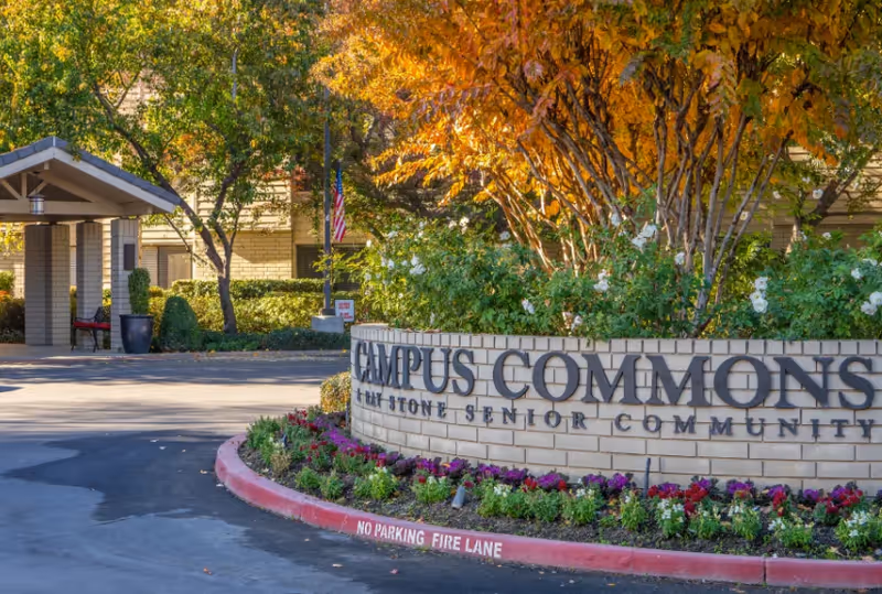 Entrance to Campus Commons Senior Living with a brick sign and landscaped flowers.