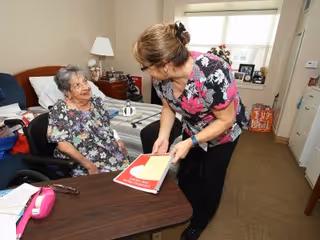 An elderly woman sitting in a wheelchair at a table in a bedroom, interacting with a caregiver who is holding a red folder. The room has a bed, a lamp, and personal items on a dresser near a window with blinds.