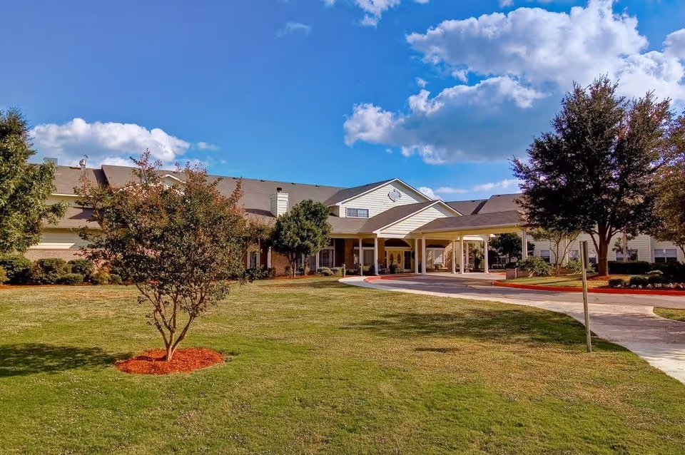 Exterior view of a senior living facility building with a covered entrance driveway, surrounded by green lawn and trees under a partly cloudy blue sky.