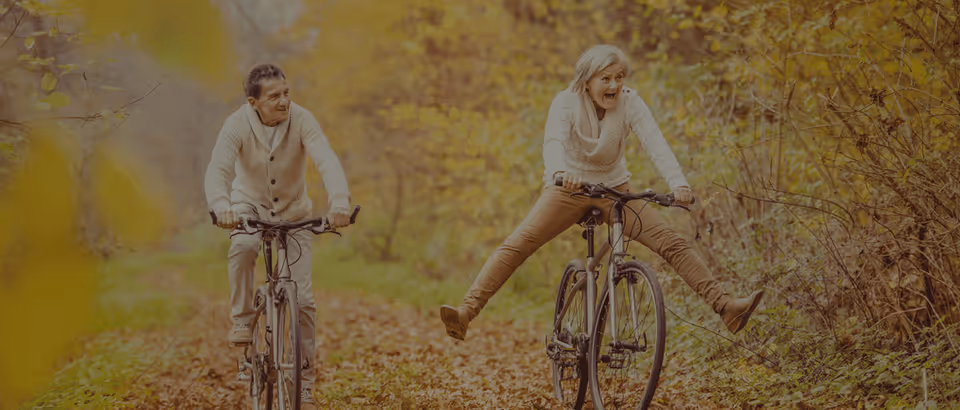 Two seniors ride bicycles along a leafy autumn forest path, smiling and enjoying outdoor activity.